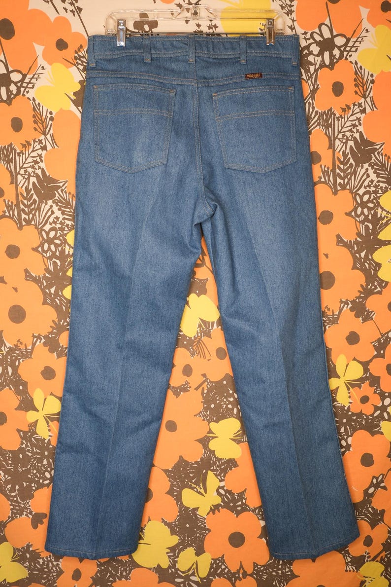 May include: Light blue denim jeans with a classic five-pocket design. The jeans have a straight leg cut and a Wrangler brand label on the back pocket. The jeans are displayed against a floral patterned background.