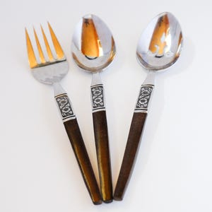May include: A set of three vintage silverware pieces: a fork, a spoon, and a slotted spoon. The fork tines are gold-colored. The spoons have a silver finish. Each piece has a dark brown handle with a decorative floral design.