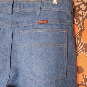 May include: Close-up of light blue denim jeans with a single back pocket. The jeans have a brown "Wrangler" label and are hanging on a clear hanger. The stitching is a contrasting tan color.