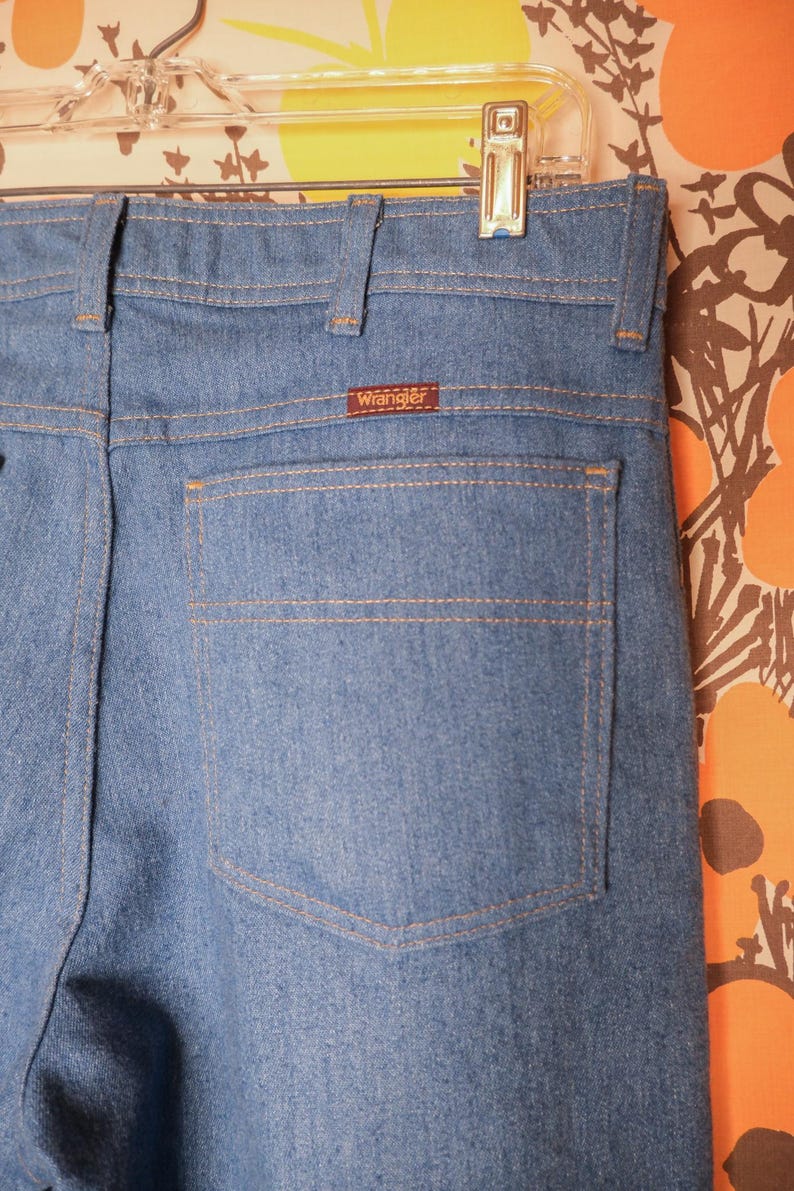 May include: Close-up of light blue denim jeans with a single back pocket. The jeans have a brown "Wrangler" label and are hanging on a clear hanger. The stitching is a contrasting tan color.