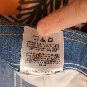 May include: Close-up of a denim jeans label with care instructions in English and Spanish. The label includes washing symbols and text indicating machine wash cold, non-chlorine bleach, tumble dry low, or line dry.