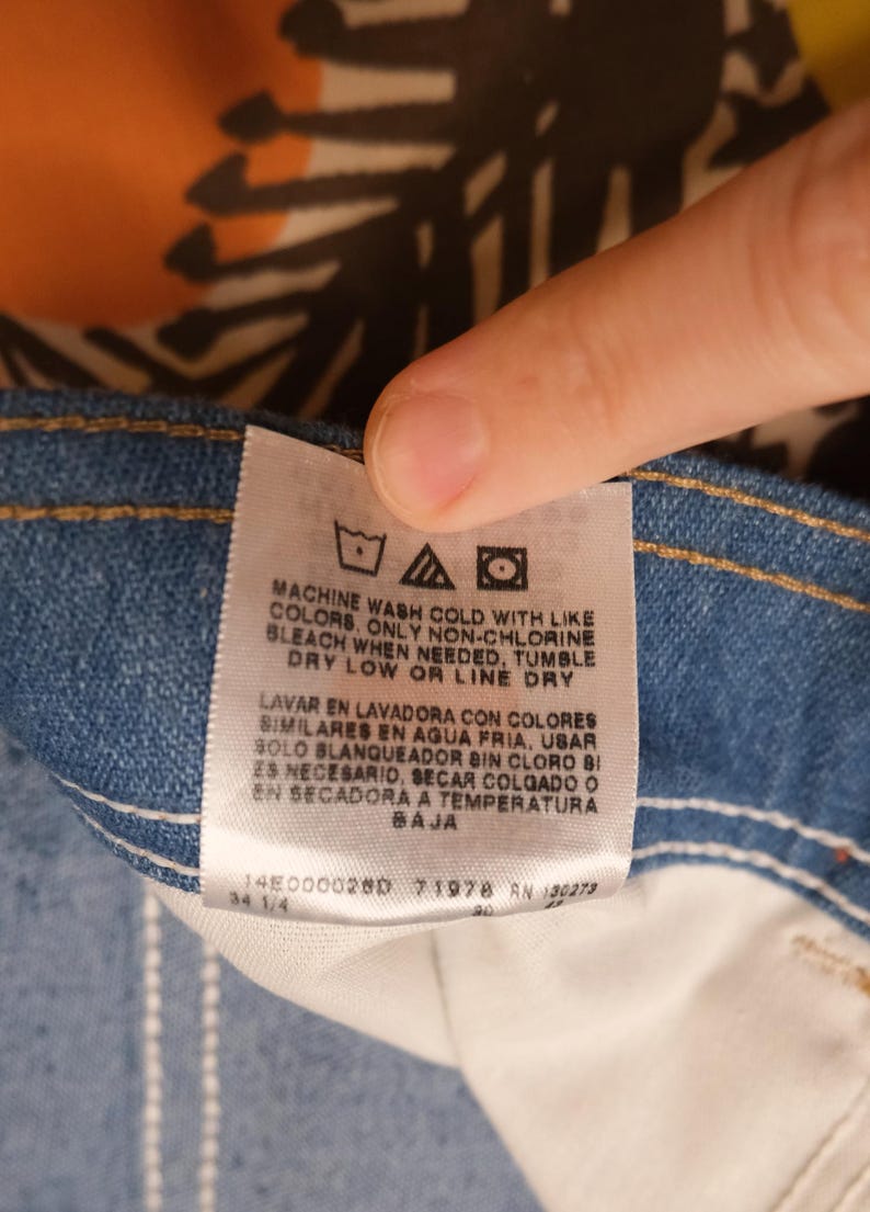 May include: Close-up of a denim jeans label with care instructions in English and Spanish. The label includes washing symbols and text indicating machine wash cold, non-chlorine bleach, tumble dry low, or line dry.