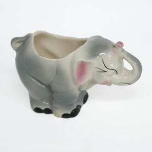 May include: A ceramic elephant-shaped planter or vessel featuring a glossy gray finish with pink accents on the ears and cheeks. The elephant has black painted eyes and toenails, with an open top cavity for holding small items or plants.