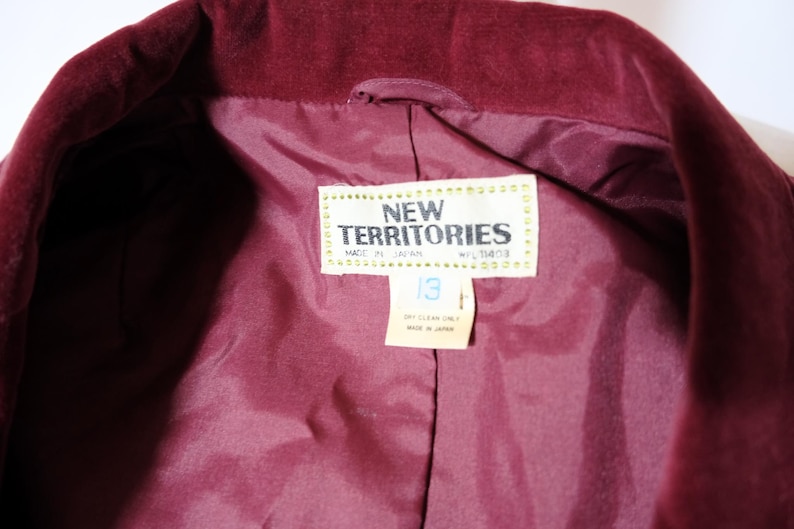 May include: Close-up of a burgundy velvet jacket interior. The lining is a matching shade of burgundy. A tag reads "NEW TERRITORIES" and "MADE IN JAPAN". The tag also indicates a size 13. Dry clean only.