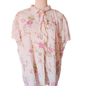 May include: A light pink short-sleeved blouse with a floral pattern of pink, purple, and orange flowers and green leaves. The blouse has a ruffled collar and a button-down front. The blouse is displayed on a mannequin.