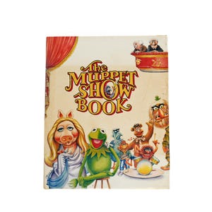May include: A hardcover book titled The Muppet Show Book featuring a colorful illustration of Miss Piggy, Kermit the Frog, Fozzie Bear, Animal, and other Muppet characters on the front cover with red stage curtains and a balcony.