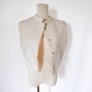 May include: A vintage, ivory-colored sequined bolero jacket with a mandarin collar and three button closure. The jacket is displayed on a mannequin against a white backdrop. The sequins shimmer, reflecting light.
