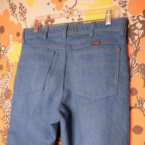 May include: Light blue denim jeans with two back pockets and a brown Wrangler label. The jeans are hanging on a clear hanger against a floral patterned background. The stitching is a contrasting tan color.