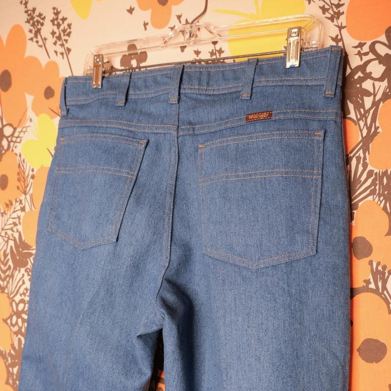 May include: Light blue denim jeans with two back pockets and a brown Wrangler label. The jeans are hanging on a clear hanger against a floral patterned background. The stitching is a contrasting tan color.