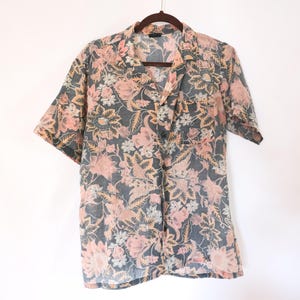 May include: Short-sleeved button-down shirt with a floral pattern. The shirt features a dark gray background with pink, peach, and white floral designs. The shirt is hanging on a wooden hanger.