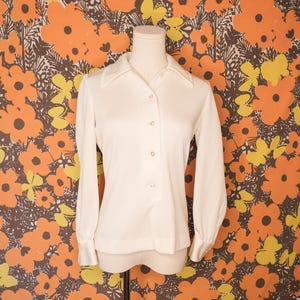 May include: A vintage cream-colored blouse with long sleeves and a collared neckline. The blouse features a button-down front and cuffs. The garment is displayed on a mannequin against a backdrop of orange, yellow, and brown floral wallpaper.