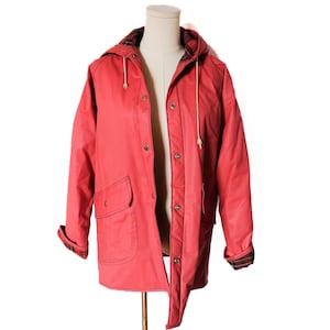 May include: A red hooded raincoat with a button-up front and two front pockets. The interior lining and cuffs feature a plaid pattern. The jacket is displayed on a mannequin against a white background. The hood has a drawstring.