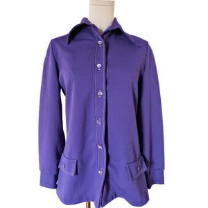 May include: A vintage purple button-down shirt with a pointed collar and long sleeves. The shirt features white stitching along the button line and around the two front pockets. The shirt is displayed on a mannequin.