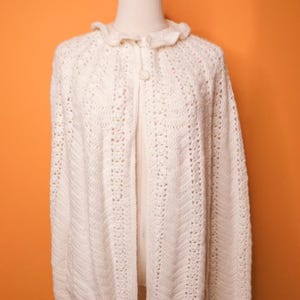 May include: Cream-colored, open-front crochet cardigan with a ruffled collar and a single button closure. The intricate openwork design creates a delicate, textured appearance. This vintage-style sweater is a cozy and stylish outerwear option.