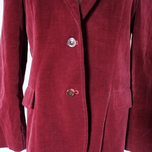 May include: A burgundy velvet blazer with two buttons and two front pockets. The blazer is displayed on a mannequin. The buttons are a silver color. The blazer is a solid color.