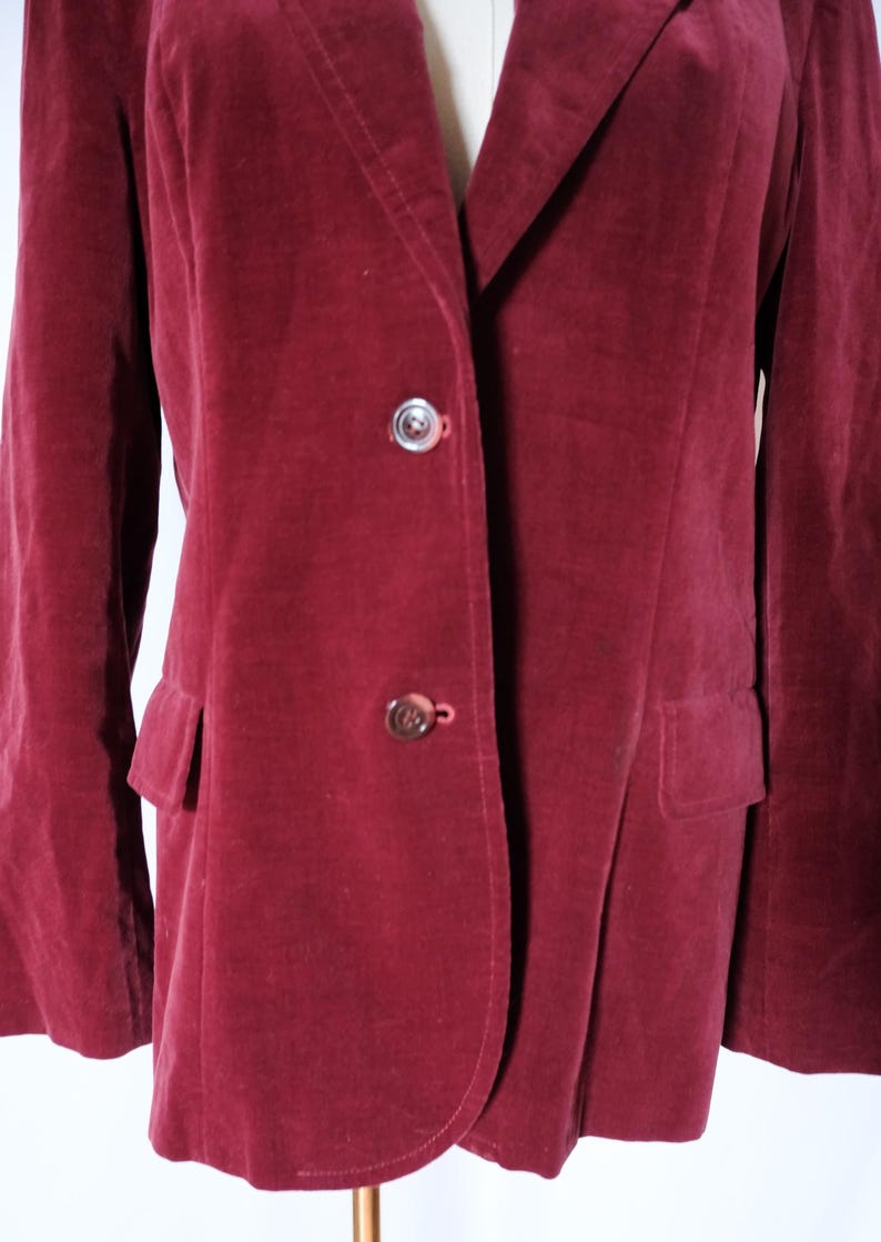 May include: A burgundy velvet blazer with two buttons and two front pockets. The blazer is displayed on a mannequin. The buttons are a silver color. The blazer is a solid color.