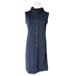 May include: A sleeveless navy blue dress with a high collar and a row of gold-colored buttons down the front. The dress is displayed on a mannequin and has a simple, elegant design. The dress appears to be made of a smooth, woven fabric.
