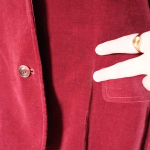 May include: A close-up of a burgundy velvet blazer. The blazer has two buttons and a pocket. The fabric has a soft texture. The blazer is a deep red color.
