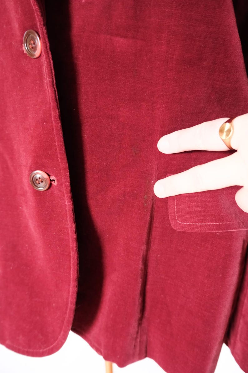 May include: A close-up of a burgundy velvet blazer. The blazer has two buttons and a pocket. The fabric has a soft texture. The blazer is a deep red color.