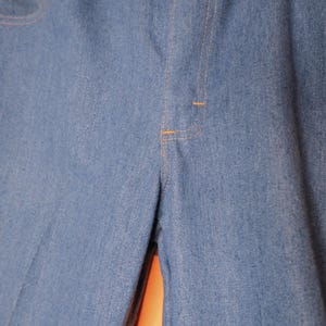 May include: Close-up of light blue denim pants. The pants have a classic design with a button and zipper closure, front pockets, and orange stitching. The fabric appears to be a medium-weight denim.