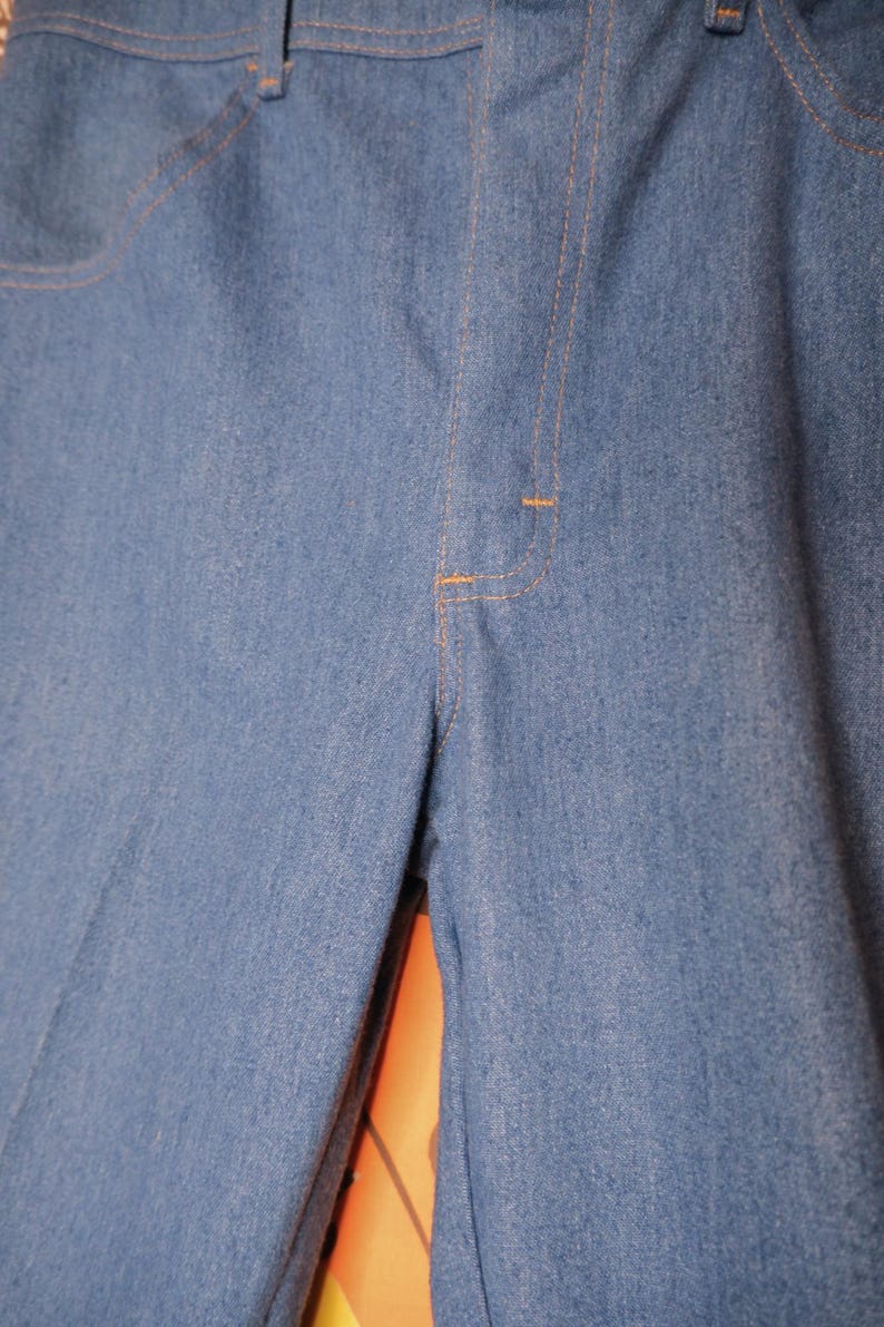 May include: Close-up of light blue denim pants. The pants have a classic design with a button and zipper closure, front pockets, and orange stitching. The fabric appears to be a medium-weight denim.