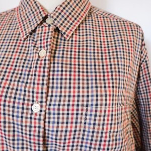 May include: A button-down shirt with a classic checkered pattern in red, blue, brown, and beige. The shirt has a collar, a chest pocket, and white buttons. The fabric appears to be a woven material.