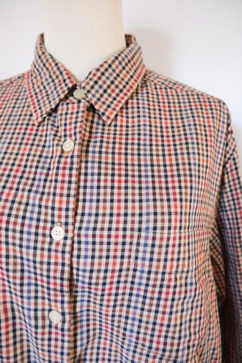 May include: A button-down shirt with a classic checkered pattern in red, blue, brown, and beige. The shirt has a collar, a chest pocket, and white buttons. The fabric appears to be a woven material.