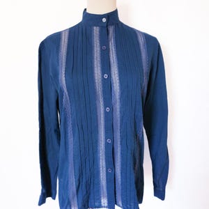 May include: A dark blue long-sleeve blouse with a high collar and button-down front. The blouse features vertical pleats and lace-like detailing down the front. The sleeves have buttoned cuffs.