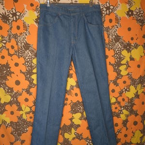 May include: A pair of light blue denim jeans hanging on a clear plastic hanger. The jeans have a classic five-pocket design and a straight leg cut. The background features a floral pattern with orange, yellow, and brown flowers.