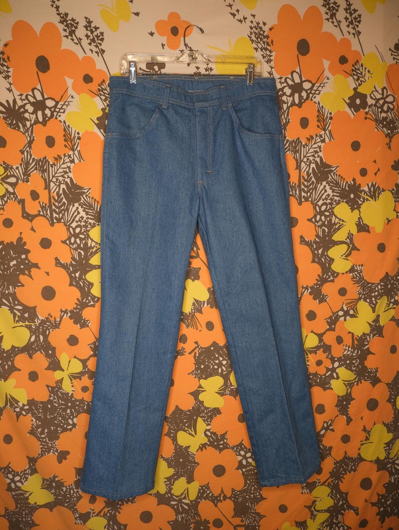 May include: A pair of light blue denim jeans hanging on a clear plastic hanger. The jeans have a classic five-pocket design and a straight leg cut. The background features a floral pattern with orange, yellow, and brown flowers.