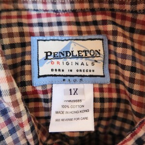 May include: Close-up of a Pendleton Originals plaid shirt label. The label reads "PENDLETON ORIGINALS BORN IN OREGON PLUS 1X." The shirt is a mix of red, blue, and brown plaid.