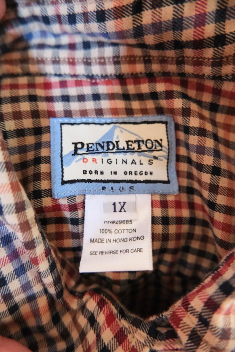 May include: Close-up of a Pendleton Originals plaid shirt label. The label reads "PENDLETON ORIGINALS BORN IN OREGON PLUS 1X." The shirt is a mix of red, blue, and brown plaid.