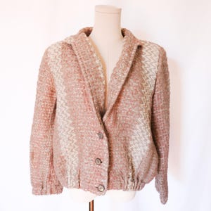 May include: A textured, button-down jacket in shades of pink and cream, displayed on a mannequin. The jacket features a unique woven pattern and a classic collar. The hemline is gathered, and the jacket has three buttons.