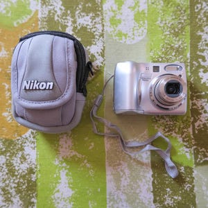 May include: A silver Nikon Coolpix 7600 digital camera with a matching gray carrying case. The camera has a lens, flash, and various buttons. The case has the Nikon logo and a zipper closure. A gray strap is attached to the camera.