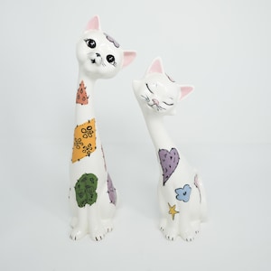 May include: Two white ceramic cat figurines with long necks and whimsical painted details. One cat has a patchwork design with orange, green, and purple accents. The other cat has a purple heart and blue flower design. Both have pink ears.