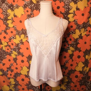 May include: A vintage white camisole with delicate lace trim and a V-neck design. The camisole features a lace panel on the bodice and a small bow detail. The garment is displayed on a mannequin against a floral patterned background.