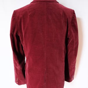 May include: A burgundy velvet blazer displayed on a mannequin. The jacket has a classic notched lapel, long sleeves with buttoned cuffs, and a single-breasted closure. The fabric has a soft, plush texture, and the color is a deep, rich red.
