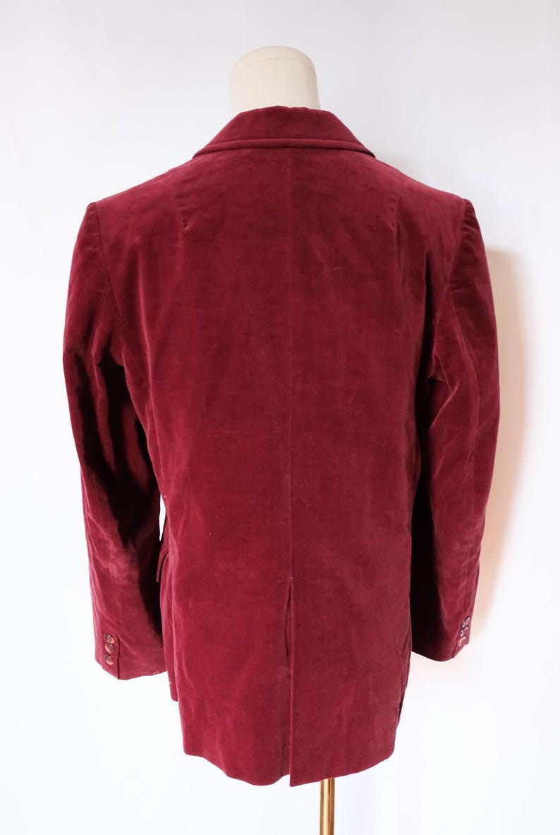 May include: A burgundy velvet blazer displayed on a mannequin. The jacket has a classic notched lapel, long sleeves with buttoned cuffs, and a single-breasted closure. The fabric has a soft, plush texture, and the color is a deep, rich red.