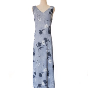 May include: A sleeveless, floor-length dress in a light blue hue, adorned with a floral pattern featuring white and navy blooms. The dress has a V-neckline and is displayed on a mannequin. The fabric appears lightweight and suitable for warmer weather.
