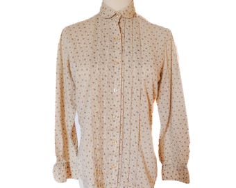 May include: A cream-colored button-down shirt with a small floral pattern. The long-sleeved blouse features a rounded collar, vertical pleats down the front, and button closures. The cuffs have a ruffled design.