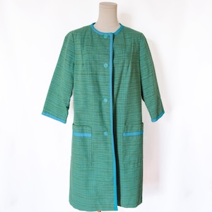 May include: A vintage green coat with three-quarter sleeves, a round neckline, and two front pockets. The coat features light blue trim along the edges, pockets, and buttons. The coat is displayed on a mannequin against a white background.