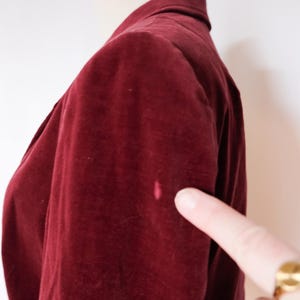 May include: A close-up of a burgundy velvet blazer on a mannequin. The blazer has a classic design with a collar and a button closure. A finger points to a small pink stain on the sleeve.