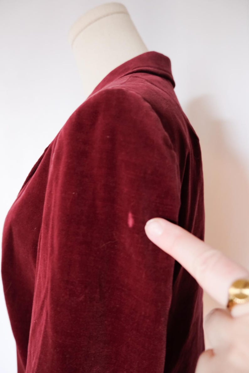 May include: A close-up of a burgundy velvet blazer on a mannequin. The blazer has a classic design with a collar and a button closure. A finger points to a small pink stain on the sleeve.
