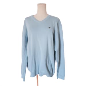 May include: A light blue long-sleeve sweater with a V-neckline and ribbed cuffs and hem. The garment features a small embroidered green crocodile logo on the left chest area. The sweater is displayed on a mannequin torso against a white background.