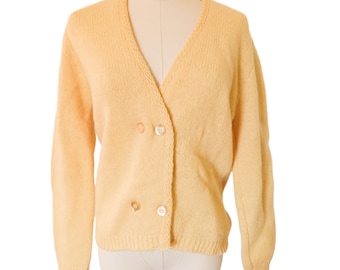 May include: A pale yellow long-sleeved cardigan featuring a V-neckline and a double-breasted front closure with four light-colored buttons. The knit garment has ribbed cuffs and a ribbed hem, providing a structured fit on the torso.