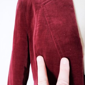 May include: Close-up of a burgundy velvet blazer. The blazer has a classic lapel design and a single-button closure. The fabric appears soft and has a subtle sheen. The image shows the texture and color of the garment.