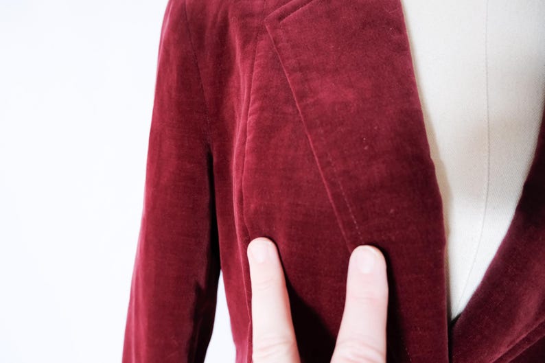 May include: Close-up of a burgundy velvet blazer. The blazer has a classic lapel design and a single-button closure. The fabric appears soft and has a subtle sheen. The image shows the texture and color of the garment.