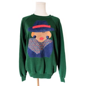 May include: A green sweatshirt featuring a graphic of a woman's face wearing a blue hat with a red band, large orange earrings, and a patterned scarf. The woman's face is tan with red lips. The sweatshirt is displayed on a white mannequin.