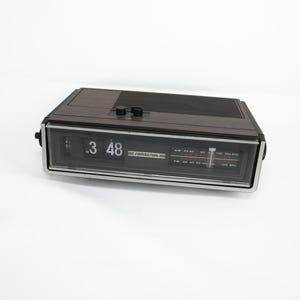 May include: A vintage FOUNDESIGN digital alarm clock with a wood-grain finish. The clock displays the time as 3:48. It has a silver-colored frame and a black speaker grill on top. The radio dial is visible below the time display.