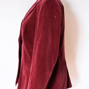 May include: A burgundy velvet blazer displayed on a mannequin. The blazer features a notched lapel, a two-button closure, and a single chest pocket. The sleeves have three buttons at the cuff. The jacket is a rich, deep red color.